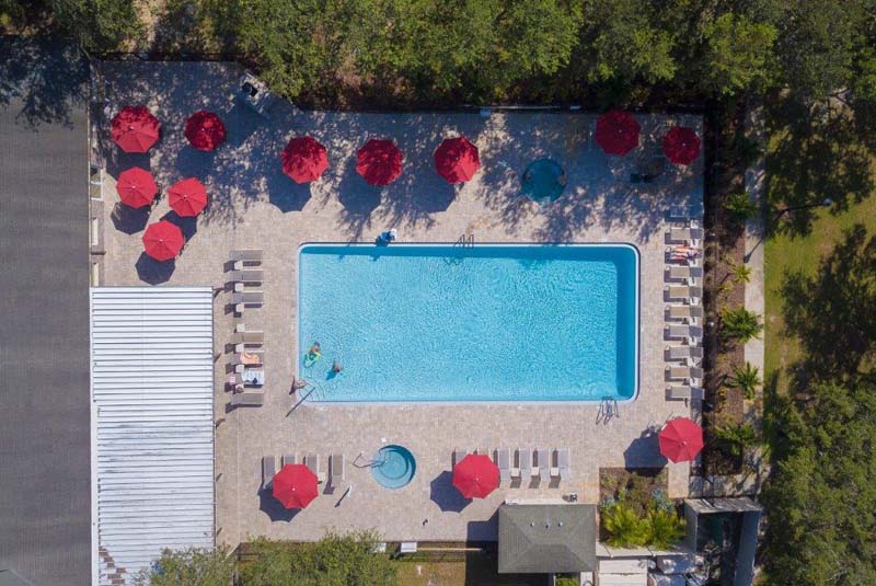Aerial view of clubhouse pool with sunning chairs and dining tables beneath red umbrellas 
